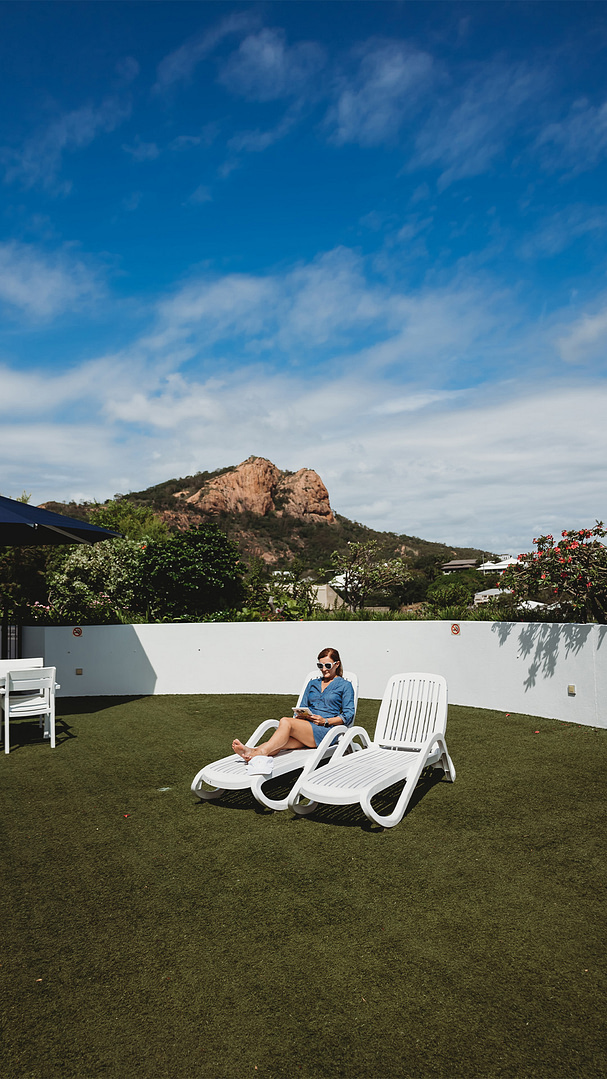 A young woman sits on a lounge chair in her yard on a blue sky, sunny day with Castle Hill in the background.